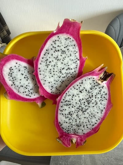 A yellow, square bowl holds three cut pieces of dragon fruit. The dragon fruit is white with black seeds and has a bright pink skin. The bowl sits in what appears to be a domestic setting, likely a kitchen or dining area, based on the presence of a grey chair visible in the background. The location is unknown but stated to be in Kraskovo, Russia.