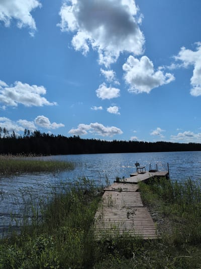 The image presents a sunlit lakeside view in Hartola, Finland, under a bright blue sky with scattered white cumulus clouds, some showing grey shadows on their undersides. A dense, dark green coniferous forest forms a horizontal band along the distant shoreline, silhouetted against the sky. The lake water is a deep blue, rippled by a light breeze, and exhibits numerous bright specular reflections from direct sunlight across its surface.
A weathered wooden pier extends from the right foreground into the lake. Positioned near the end of the pier is a single, dark-toned wooden chair, facing away from the viewer towards the open water. Adjacent to the chair, a set of metal steps or rails for water entry is visible at the pier's edge. The foreground consists of lush green vegetation, including tall reeds and grasses growing along the water's edge and bordering the wooden pathway leading to the pier. Small yellow wildflowers are interspersed within the foreground greenery. The scene is devoid of human or animal presence.