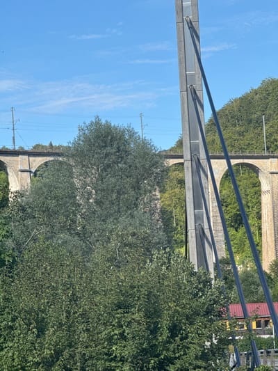 The image depicts a rural landscape in Clos du Doubs, Switzerland, featuring a large, arched stone railway viaduct in the background, partially obscured by dense green foliage. The viaduct, constructed of light-colored stone, has multiple arches spanning across a valley. Above the viaduct, power lines and railway tracks are visible. In the foreground, a tall, modern concrete pillar with dark cables attached stands prominently, suggesting it is part of a newer infrastructure project that may be integrated with or adjacent to the historic viaduct. The pillar is segmented and has a vertical light panel in its center. A lush, green hillside covered in trees rises behind the viaduct, indicating a natural, forested environment. In the lower right quadrant of the image, a building with a red roof and yellow siding is partially visible, hinting at a nearby settlement or industrial area. The sky is a clear, bright blue with scattered wispy white clouds, indicating fair weather. The overall scene captures a juxtaposition of historical and modern infrastructure within a natural Swiss setting.