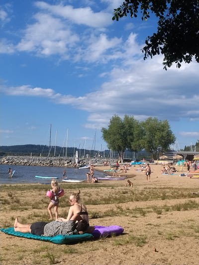The image depicts a sunny day at a beach, likely in Radslav, Czechia, evidenced by the presence of a lake or reservoir with sailboats in the background and a rocky breakwater. In the foreground, two individuals are lying on an inflatable mattress on the sand. A woman with blonde hair pulled back in a bun, wearing sunglasses and a black bikini top, is sitting up and looking to her right, with her hand on the shoulder of the person lying down. The person lying down has dark hair and is wearing black shorts and a patterned shirt. A young child wearing pink arm floaties and a swimsuit is walking away from them towards the water. Further down the beach, numerous people are scattered, some sunbathing on towels, others walking, and several are in the water, some with paddleboards. A cluster of trees is visible in the middle ground, with a shoreline and forested hills in the distance. The sky is blue with scattered white clouds.