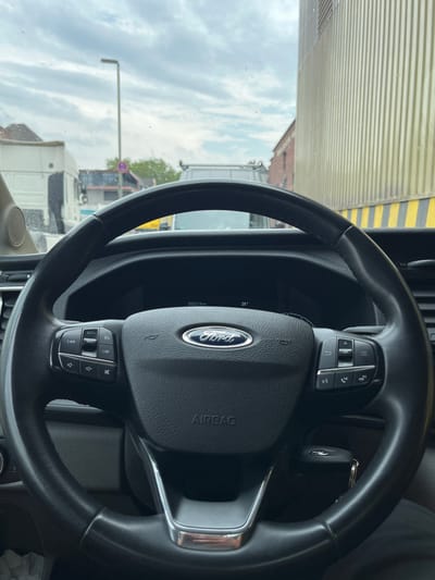 The image shows the interior of a vehicle, likely a van or truck, as seen from the driver's seat. The focus is on the steering wheel and dashboard. The steering wheel is black, with a Ford logo prominently displayed in the center. Various buttons are visible on the steering wheel, including controls for cruise control, volume, and phone functions. A car key with a Ford emblem is hanging from the ignition.
Through the windshield, an outdoor scene is visible, though somewhat distorted by the glass. There are buildings with brickwork and a cloudy sky. A traffic sign, possibly indicating a no parking zone, is visible in the distance. The overall lighting suggests it is daytime.
In the background, through the side window or reflection, parts of what appear to be other vehicles and industrial structures can be seen, including a large corrugated metal wall with yellow and black stripes. The presence of a truck or van interior, along with the outdoor view, suggests the vehicle is either parked or in transit. No people are visible in the image. The location context is Neuss, Germany, which aligns with the visual cues.