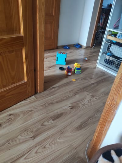 The image depicts a domestic interior scene. A wooden door is partially open on the left, revealing a hallway with light brown wood-patterned laminate flooring. In the middle ground of the room, a bright blue plastic toy, possibly a tablet or interactive toy, is on the floor, along with two small toy vehicles: a red and yellow truck and a green and yellow truck. Further to the right, a white shelf unit stands against the wall, containing various items including a white wire basket with clothing or fabric. Two blue children's sandals are on the floor near the base of the wall on the left. The environment appears to be a child's play area or a general living space. There are no people visible in the image. The scene is consistent with a residential setting. The location is identified as Oława, Poland, though this information is not visually discernible from the image itself.