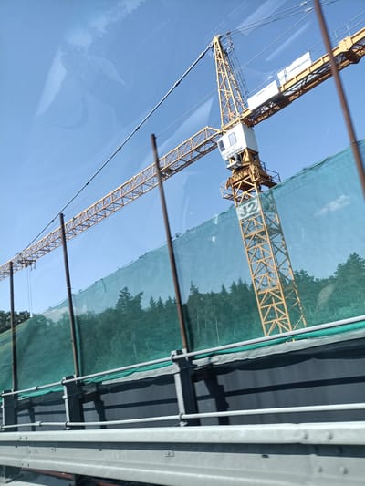 A yellow tower crane is visible against a bright blue sky, angled from the lower right to the upper left of the frame. The crane has a cabin with "LIEBHERR" visible on its side. A large green construction net is stretched across the middle ground, obscuring the lower portion of the crane's mast and the trees behind it. The net is supported by vertical poles. In the foreground, there is a grey guardrail. The scene appears to be part of a construction site, possibly located in Hrusice, Czechia, given the metadata. There are no people visible in the image. The overall lighting suggests it is daytime.
