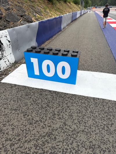 A slightly angled shot showcases a racetrack section. A blue sign with the number "100" in white is prominently displayed next to a solid white line. The track's surface has varying textures; the right side appears more rough and granular, while the left features smoother asphalt with painted colors. A person walks distantly in the top left of the frame. The side of the track features a painted blue and silver border.