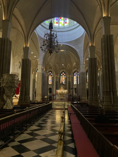 The image displays the interior of a grand cathedral or church, looking down a long central nave towards the distant altar area. The perspective highlights the impressive scale and architectural complexity of the building.
The ceiling features high, light-colored ribbed vaulting, characteristic of Gothic or Neo-Gothic architecture, supported by substantial, fluted columns lining both sides of the nave. Multiple ornate, dark metal chandeliers with many lights hang from the ceiling, contributing to the interior illumination.
Vibrant stained glass windows are visible on the left side, some featuring abstract geometric patterns in bright colors, while others depict detailed figures or scenes. A brightly lit area further down the nave, illuminated by more stained glass, appears to be the main altar or chancel, adorned with religious artifacts and candles.
The floor of the central aisle boasts a distinctive black and white checkered pattern. To the right, a long row of dark wooden pews with plush red cushions stretches along a side aisle, separated from the main walkway by a decorative black metal railing topped with gleaming brass finials. In the left foreground, a richly carved stone or marble sculpture base, possibly part of a pulpit or a monument, adds to the decorative elements.
The scene conveys a sense of solemn grandeur and historical depth. The building appears to be empty of people, suggesting a quiet moment. The lighting is a blend of natural light filtering through the stained glass and artificial illumination from the chandeliers and other fixtures, creating a serene and contemplative atmosphere. No visible text is present in the image.