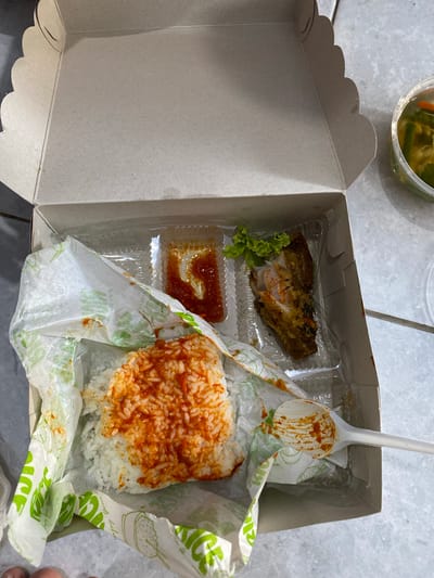The image shows a white cardboard takeout box, open and placed on a light-colored, possibly tiled surface. Inside the box, there are three main food items: a portion of white rice topped with a red sauce, a small container of red sauce, and a piece of fried chicken with a small garnish of lettuce. A white plastic spoon with traces of sauce rests on the edge of the box, near the rice. To the right of the box, a small, clear plastic container holds pickles or pickled vegetables. The overall scene suggests a meal that has been opened for consumption. The environment appears to be indoors, with diffused lighting. There is no clear indication of the specific location within Madiun, Indonesia, beyond it being a place where such food would be served or consumed.