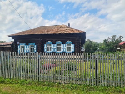 The image depicts a rustic wooden house with blue shutters, typical of rural Russian architecture, in Малая Белоносова, Russia. The house is constructed from dark logs and has a weathered, corrugated metal roof. Three windows on the visible facade are adorned with intricately carved blue and white shutters. A simple wooden fence, also showing signs of age with peeling paint, stands in front of the house, separating it from a grassy yard and flowerbeds. Lush green grass covers the foreground, with some wildflowers adding splashes of color behind the fence. In the background, dense trees form a natural backdrop, and the sky is a bright blue with scattered white clouds, suggesting it is daytime with pleasant weather. The scene evokes a sense of tranquility and traditional rural life. No people are visible in the image, and there is no discernible text.