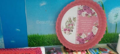 The image shows a pink decorative plate with a floral pattern and a quote. The plate is placed in front of a backdrop that depicts a blue sky with dandelions, green grass, and a blurry white dandelion. The quote on the plate reads: "waite for the one who is constantly reminding you how he cares a bout you & how much lucky he's to have YOU". There are also some blurry items in the foreground, including what appears to be a red textured object and some colorful patterns. There are no people or animals visible in the image. The location is not discernible from the image content itself.