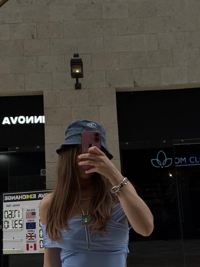 A young woman is taking a selfie in a storefront window reflection in Ciudad López Mateos, Mexico. She is wearing a blue denim bucket hat, a blue top, a silver bracelet, and a bolo tie-style necklace. She is holding a pink smartphone. Behind her, the reflection shows an Avon store and an exchange rate board displaying rates for USD, EUR, GBP, and CAD. A wall with a mounted light fixture is also visible in the reflection.