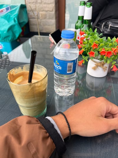 The image depicts a close-up, first-person perspective of a table setting at a cafe in Djigurovo, Bulgaria. In the foreground, the bare forearm and hand of a person, exhibiting a light skin tone, rest on a dark, reflective table surface. The individual wears a black beaded bracelet and a grey wristband, with the cuff of a brown jacket visible.
On the table, a clear glass contains a layered beverage, featuring a translucent light-green liquid at the bottom and a frothy, light-brown substance, consistent with a coffee or frappe, on top. A black straw is inserted into this drink. To its right, a plastic bottle of "Nazlı Spring Water" with a blue cap stands upright, displaying a barcode on its label. Further to the right, a small white ceramic pot, adorned with a simple twine bow, contains artificial red-orange bell-shaped flowers and green foliage.
In the blurred background, several items are visible on the same table: two green glass bottles, appearing to be beer bottles with white labels, and a metallic can. A black smartphone with a dark screen lies on the table near a partially visible hand of another individual. Beyond the table, a large, light-blue object with white lettering is partially visible on the left, while a light-colored building facade and a slender, light-toned vertical object are present in the center. A black bag is also discernible in the upper right background.