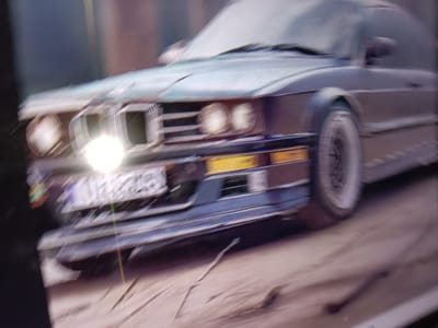 The image shows a blurred close-up of the front of a dark-colored sedan parked on a sandy or dirt surface. The car has its headlights on, one of which is very bright and causes lens flare. The front grille is visible, along with the bumper which has yellow indicators and a license plate. The car appears to be a classic model, possibly from the 1980s or 1990s, with sharp, angular lines. The background is out of focus and appears to be a dark, indistinct area. There is no visible signage or landmarks to indicate a specific location within Alessandria, Italy.
