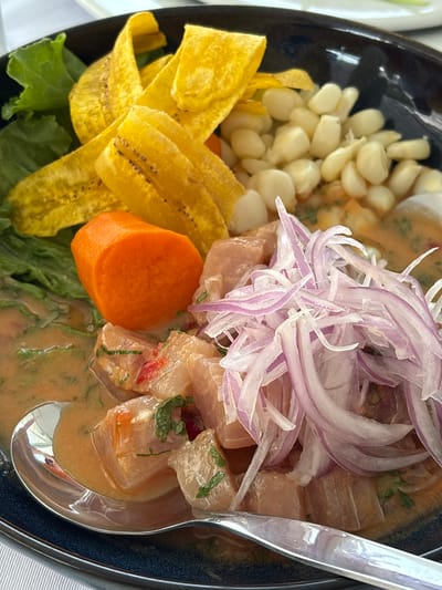This is a close-up, eye-level, outdoor, daylight, medium shot of a bowl of Peruvian ceviche. The ceviche is served in a dark blue bowl and consists of chunks of white fish marinated in a light orange sauce. It is topped with thinly sliced red onions, a segment of cooked sweet potato, corn kernels, and plantain chips. There is also a sprig of lettuce visible. A silver spoon rests in the sauce, partially submerged. The background is blurred, but suggests an outdoor dining setting with hints of white tablecloths. The dish is likely prepared in La Punta, Peru, a district known for its seafood restaurants.
