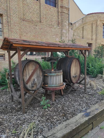 In the city of Brühl, Germany, an outdoor scene depicts a rustic wine press and two large wooden barrels situated beneath a wooden shelter with a tiled roof. The shelter is supported by dark wooden beams, and the ground is covered with wood chips. In the background, a building constructed of light brown bricks with multiple windows is visible, suggesting an industrial or historical setting. Lush green bushes and some dry foliage surround the wine-making equipment, contributing to a natural and somewhat aged atmosphere. The lighting suggests it is daytime, possibly late afternoon due to the soft light. No people are present in the image.