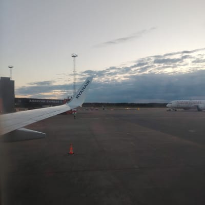 The image captures an airport tarmac scene at Stockholm-Arlanda, Sweden, viewed from a low perspective, likely an aircraft window, during either dawn or dusk. In the left foreground, the white wing of a Ryanair aircraft is prominent, with "RYANAIR" visible in blue on the winglet. An orange traffic cone rests on the dark gray asphalt tarmac beneath the wing.
In the left midground, a dark jet bridge is visible, marked with "18" and "AMERICAN EXPRESS" in white text. To the far left, a dark building structure and a tall metal lattice tower with a circular platform are present. A single ground crew member in a high-visibility yellow-green vest stands on the tarmac near the Ryanair wing.
Towards the right midground, the forward fuselage and nose of an Ethiopian Airlines aircraft are discernible, with "Ethiopian" clearly legible on its side. A pushback tug is connected to the nose landing gear of the Ethiopian aircraft. In the distance, several small baggage carts are scattered across the tarmac. Another tall metal lattice tower with a platform is visible in the background.
A dark line of trees or forest defines the horizon beyond the tarmac. The sky is partially cloudy, with diffused light indicating a low sun angle consistent with either sunrise or sunset.