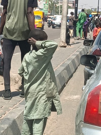 The image depicts an urban street scene in Hotoro, Nigeria, under bright sunlight. In the foreground, a young boy, dressed in a light green long-sleeved tunic and matching trousers, stands facing partially away from the viewer, his right arm raised with his hand covering his eyes. To his immediate left, the lower half of an adult male is visible, wearing a green shirt and dark trousers, standing on a grey stone-paved sidewalk. On the far right, the rear section of a silver car is present, with its red taillight and bumper visible. Adjacent to the silver car, a black vehicle's side mirror is seen.
A striped concrete curb separates the paved sidewalk from a busy road. Along the sidewalk in the mid-ground, a black utility pole marked with the numeral "2" is visible. Several other individuals are standing or walking further down the sidewalk, including one person in a bright green outfit and another in light brown. On the street beyond the curb, traffic includes a yellow compact truck or tuk-tuk, a white van, and a motorcycle with a rider. The background features distant buildings, green trees, and large commercial signage with green and white elements. Strong shadows indicate a high sun.