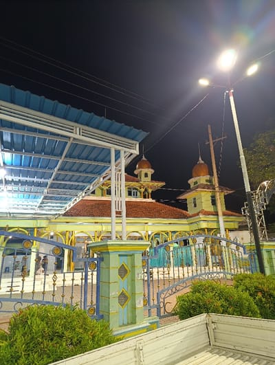 The image shows a mosque in Kecamatan Kapongan, Indonesia (latitude: -7.675, longitude: 114.065). The mosque has a green and yellow facade, with a red roof. The front of the mosque is enclosed by a blue and gold gate. Two people walk within the mosque. Two domes are visible on the roof, with a golden design. A blue metal awning is extending out from the mosque, covering a portion of the sidewalk. A streetlamp is shining brightly overhead, with light reflecting off the wet pavement. A white truck bed is positioned in the foreground with a bushy green plant to the left of the truck bed.  The sky is dark blue, with the light from the streetlamp casting a bright glare.
