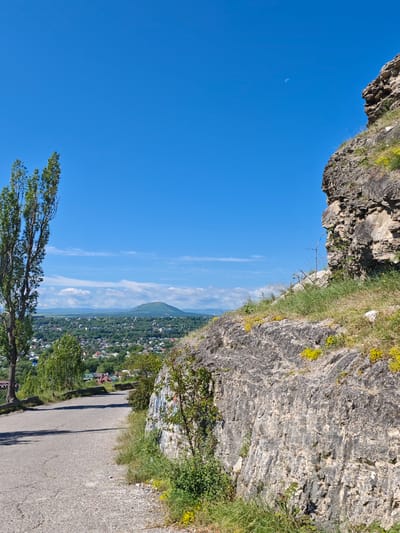 The image captures a scenic view from an elevated position overlooking the city of Goryachevodsky, Russia. In the foreground, a paved road curves upwards, bordered by a rugged, rocky embankment on the right. The embankment is sparsely covered with green grass and small yellow wildflowers. A tall, slender tree stands to the left of the road, its foliage dense and green.

Beyond the road and tree, the city of Goryachevodsky sprawls across a verdant valley. Numerous buildings with red and white roofs are nestled among lush green trees. In the distance, a prominent, conical mountain rises against a clear, bright blue sky. Wispy white clouds dot the upper portion of the sky. A faint crescent moon is visible in the upper right quadrant of the sky. The overall impression is one of a tranquil, sun-drenched day in a picturesque Russian locale.