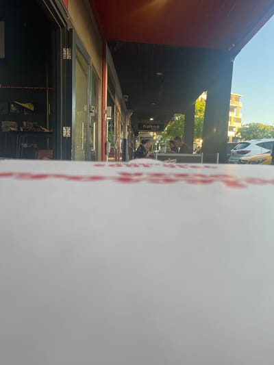 The image depicts a street scene in Applecross, Australia, specifically on a commercial strip, likely in front of "Haiven Espresso & Deli". The perspective is low-angle, looking over a blurred white surface with faint red patterns in the foreground, possibly a tabletop or a container lid.
In the midground, a pedestrian walkway is visible, partially shaded by a dark awning extending from a row of commercial buildings. A sign displaying "haiven" in white text is mounted under this awning. To the left, a storefront with dark walls and large, partially open glass bi-fold doors is visible.
Two individuals are seated at an outdoor table under the "haiven" awning. The person on the left, appearing to be female, has dark hair and is wearing a dark garment, looking down at an object on the table. The person on the right, appearing to be male, has short dark hair and is wearing a dark shirt, facing the first individual.
In the background, to the right of the seated individuals, a light-colored SUV is parked on the street. Beyond the street, a multi-story building with balconies is visible under a clear blue sky, alongside green foliage from trees. The scene is illuminated by natural light, indicating daytime.