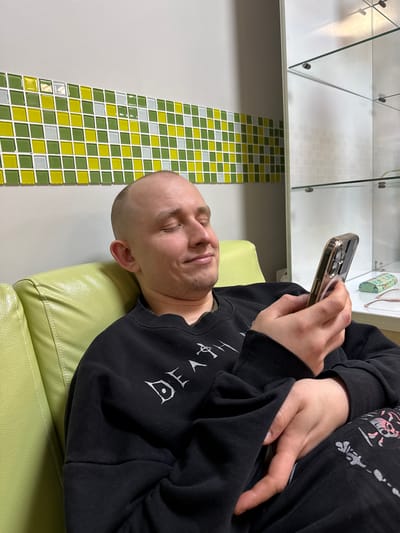 A man with a shaved head is reclining on a light green upholstered bench or couch, holding a cell phone in his right hand and looking at the screen. He is wearing a black sweatshirt with a graphic design on the front. The area has a section of wall with small, square tiles in shades of green, yellow, and white. Above that is a white wall and a glass shelving unit or display case. The location is in an indoor area, likely in the city of Kuleshovka, Russia.
