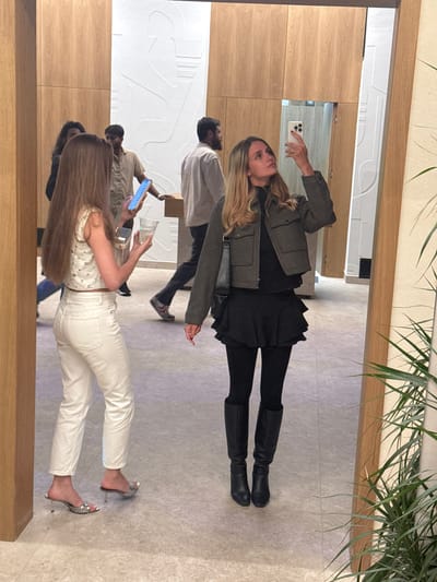 In Akhalubani, Georgia, a woman with blonde hair is captured in a mirror reflection, taking a selfie with her phone. She is wearing a dark turtleneck, a grey cropped jacket, a black ruffled skirt, black tights, and black knee-high boots. She is also carrying a black purse over her shoulder. In the foreground, another woman with long blonde hair, wearing a white sparkly top and white pants, is holding a phone and a drink. Other individuals are visible in the background. The scene appears to be indoors, possibly in a public space with wooden paneling and a white textured wall.
