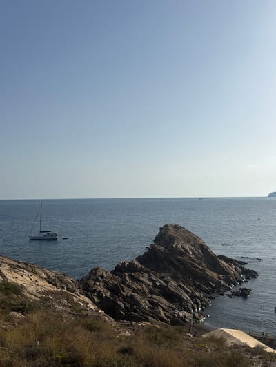 The image depicts a coastal scene in La Unión, Spain, characterized by a clear sky and calm sea. In the foreground, a rocky outcrop covered in dry grass and brush slopes down towards the water. A large, jagged rock formation juts out into the sea, its dark, striated surface contrasting with the blue water.
Further out in the sea, a white sailboat with a single mast is visible, appearing to be at anchor. A small inflatable dinghy is tied to the stern of the sailboat. A few other distant boats can be seen on the horizon.
On a small sandy beach nestled between rocks, a few people are visible. One person appears to be standing on the beach, and another is further down, near a light-colored sunshade or awning. Several swimmers are dotted in the water near the rocky shore. The overall atmosphere is one of peaceful coastal recreation.