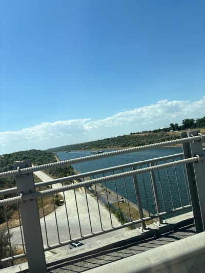 The image shows a view from a bridge overlooking a canal in Agigea, Romania. The canal is wide and lined with green, tree-covered hills on both sides. Several barges are visible on the water. The foreground of the image is dominated by a gray metal railing with thick ropes running horizontally through it. Below the railing, a concrete path or road is visible, with a light-colored car parked on it. The sky is a clear blue with a few scattered white clouds. The overall impression is one of a bright, sunny day in an industrial or maritime setting.