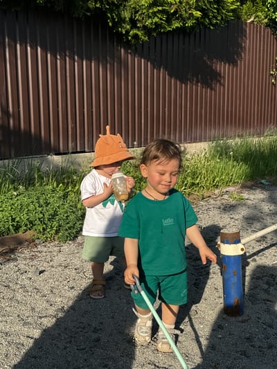 In Селекционер-2 Снт, Russia, two children are outdoors on a sunny day. One child, wearing a white shirt, green shorts, and a brown hat with antennae, holds a bottle. The other child is dressed in a green shirt and green shorts, and holds a fishing rod. They are standing on gravel near a metal fence and vegetation. A blue and white metal object is also visible in the background.