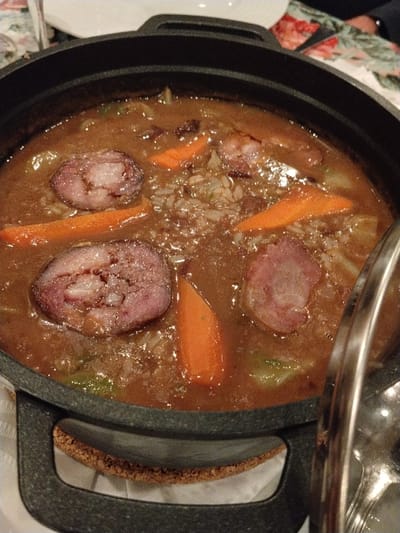 A cast-iron pot filled with a savory stew. Circular sausage slices and bright orange carrot pieces float in a rich, brown broth. Wisps of cabbage and scattered grains of rice are visible within the stew. A metal lid rests askew to the side, revealing the contents. The pot sits on a white cloth, contrasting with the dark, textured exterior of the pot.