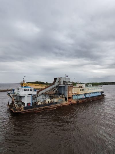 The image shows a large, weathered dredger barge, possibly stationary, in what appears to be a body of water in Ozernoe Ust'e, Russia. The barge is multi-level with a main structure predominantly white, blue and cream-colored, including the lettering "СЛАВУТИЧ" on its side. Industrial equipment such as pipes, winches, and a large chute are visible on the barge. The hull shows significant rust. The water is dark and choppy. In the background, a sandy area with green vegetation is seen, along with another boat. The sky is overcast with grey clouds.