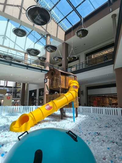 This media file depicts an indoor children's play area within a shopping mall, likely in Montijo, Portugal, given the context. The scene is dominated by a large, white ball pit filled with thousands of white, light blue, and pink balls. In the foreground, a portion of a large, blue inflatable is visible, suggesting it's part of the play equipment.
Rising from the ball pit is a wooden play structure resembling a small fort or treehouse, complete with a red-and-white skull and crossbones flag. Attached to this structure is a bright yellow, winding slide with red accents. The slide is designed as a tube, with a wide opening at the top and a gentle slope leading into the ball pit.
Above the play area, the mall's architecture is visible, featuring multiple levels with railings and shops. The ceiling is a transparent, segmented roof, allowing natural light to filter in, indicating it is daytime. Several large, dark, bowl-shaped light fixtures are suspended from the ceiling by thin wires, adding to the modern interior design.
Shops are visible on the upper levels, with one clearly displaying the text "STRADIVARIUS" in illuminated lettering. Other shop fronts have visible signage, including "JD" and what appear to be retail displays with percentages or sale signs. Although people are present in the background, they are not the main focus and appear to be shoppers or mall staff. The overall impression is a bright, safe, and engaging environment designed for children's entertainment. The presence of the play area suggests a family-friendly shopping destination.