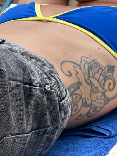 A close-up reveals a person's upper thigh and side. The skin bears a prominent black-inked tattoo of a floral design. A blue swimsuit with yellow trim peeks into the frame, contrasting with the dark gray denim shorts covering part of the thigh. A blue towel is also visible in the top right corner. The image is brightly lit and focuses on texture and color contrast.