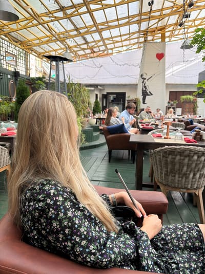 In a covered outdoor seating area of a restaurant in Zhukovka, Russia, a blonde woman is seated in a brown leather armchair, her back to the camera. She is wearing a black dress with a floral pattern. Her hands are resting on her lap, and she is holding a metal pipe. Several tables with red napkins and glasses are visible in the background, along with other diners. A large banner with a stencil of a girl holding a red balloon hangs on a white wall. The area is furnished with wicker chairs and wooden tables. Green plants and a patio heater are also present.