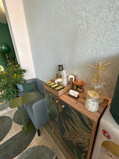 In a dimly lit, indoor commercial space, likely a cafe or waiting area, a corner is furnished with a grey L-shaped couch and a small, green, oval-shaped table. Adjacent to the couch is a wooden cabinet with a dark green, botanical-themed design. On the cabinet's surface, there are several items, including a display of individually wrapped tea bags, stacks of clear plastic cups, and a wooden rack holding clear, ribbed glasses. A small glass jar filled with individually wrapped candies sits next to a business card that reads "КОФЕРУМ" (KOFERUM) in Cyrillic, resting on a wooden stand. Another glass jar contains sugar cubes. The wall behind the cabinet has a textured, light grey finish. To the left of the couch, a large potted plant with lush green foliage is positioned near a white wall. A green balloon is partially visible behind the plant. The floor is covered by a patterned rug with large, abstract green shapes on a neutral background. A white water dispenser is partially visible to the right of the cabinet. The scene appears to be located in Medvedevo, Russia.