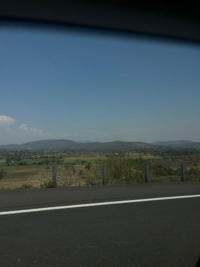 The image presents a view from a moving vehicle, likely a car or bus, traveling on a road near Tlaquiltenango, Mexico. The foreground shows a portion of the vehicle's interior on the left and the road surface on the right. A white line divides the lanes. Beyond the road, the landscape features a mix of agricultural fields, scattered trees, and small settlements situated at the base of hills. In the distance, a range of mountains is visible under a partly cloudy sky. The overall impression is one of a rural landscape bordering an urban area, viewed from a roadside perspective.