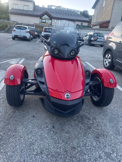 The image displays a red three-wheeled vehicle, possibly a Can-Am Spyder, parked in an outdoor lot in Lavans-lès-Saint-Claude, France. The vehicle is positioned front and center, with its bright red bodywork and black accents prominently visible. In the background, several cars are parked, including a white Renault Clio with a French license plate. Behind the parked vehicles, there is a building with a sloping roof and what appears to be a signage indicating an event, possibly a Salle des Fêtes. The environment suggests a public parking area, likely adjacent to a community building, under a clear sky with soft lighting, indicating it is either early morning or late afternoon. The ground is paved with asphalt.