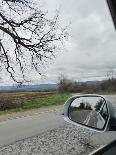 The image shows a landscape taken from inside a moving car. In the foreground, the car's side mirror reflects the road behind. The road has two white lines painted on it. Further back, there is a field, some bushes, and a range of distant hills. Above, there is a cloudy sky and the bare branches of a tree. The location is identified as Mramoren, Bulgaria.
