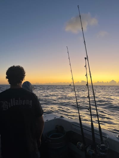 The image depicts an offshore marine scene during either sunrise or sunset in the Pacific Ocean, within the area of Wollongong City Council, Australia. The sky transitions from vibrant orange and yellow hues at the horizon to purple and blue overhead, with a single white cloud visible in the upper right quadrant.
In the foreground, the stern section of a white fishing boat with grey trim is visible. Two individuals are positioned on the boat, facing the horizon. The primary subject, an adult male, is viewed from behind, wearing a black t-shirt with the word "Billabong" in a stylized white font on the back. He has dark, curly hair. Partially obscured by this individual, the top of another person's head, covered by a light-colored hat or hood, is visible.
Three heavy-duty fishing rods with attached reels are mounted vertically along the right side of the boat, with fishing lines extending from them. A grey bucket with a blue rim is situated on the boat's deck to the left of the main subject. The water exhibits a moderate chop, reflecting the warm light from the low sun. Along the distant horizon, a low bank of clouds is present, and two distinct, smaller vessels are visible as dark silhouettes against the bright sky.