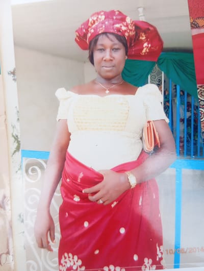 A medium-shot image shows a woman in traditional attire standing outdoors in Karsana 2, Nigeria, with a date stamp "10/05/2014" visible in the lower right corner. She is wearing a red and gold patterned headwrap, a cream-colored off-the-shoulder top with intricate lace detailing, and a vibrant red skirt embellished with gold floral patterns. The woman has a gold necklace with a small pendant and a gold wristwatch on her left wrist. She is holding a small orange clutch bag in her right hand, which is tucked under her left arm. The background features a light-colored wall, a blue railing, and green draped fabric. The overall scene suggests a festive or formal occasion.
