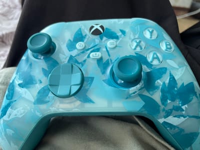 An Xbox controller with a blue and white abstract pattern is placed on a grey fabric surface, possibly a couch or bed. The controller is angled slightly towards the viewer and is the primary subject. The background is dark, with a hint of a black garment and white bedding visible. There are no people, animals, or any other objects present. The lighting appears to be soft and natural. The image provides a close-up view of the controller. The location in Lipcani, Moldova is not discernable from the image.