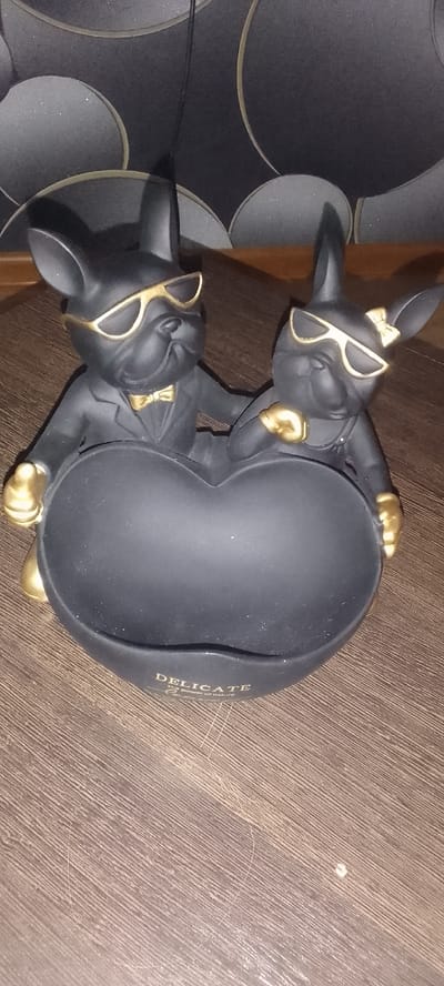 A black, heart-shaped dish is held by two black bulldog figurines wearing gold sunglasses and gold accents. The bulldog on the left is wearing a gold bowtie and a black suit jacket, while the bulldog on the right is wearing a gold ribbon on its head and overalls. Both figurines have their paws on the edge of the dish. The dish has the word "DELICATE" and "The Power of nature" inscribed on it in gold lettering. The background consists of a dark, textured wallpaper with circular patterns. The scene is set on a wooden surface. The location is specified as Topar, Kazakhstan.