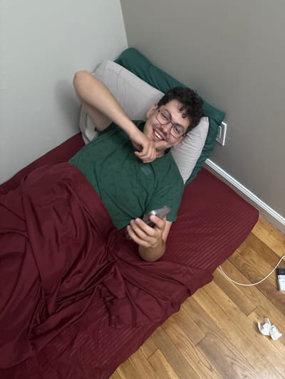 A young man, likely in his late teens or early twenties, is pictured lying on a bed in an indoor setting within Franklin Township, United States of America. He has short, curly dark hair and wears glasses along with a dark green patterned t-shirt. He is smiling broadly, looking directly at the camera, conveying a cheerful and relaxed demeanor.
He is propped up against a light grey wall, resting on a dark green pillow and a light grey/white pillow. His lower body is covered by a maroon or dark red sheet or comforter. In his right hand, he holds a grey smartphone.
The floor adjacent to the bed is made of light-colored wood planks. Visible on the wooden floor is a white charging cable connected to a white wall adapter, which has partially legible text resembling "XIAOMI." A small, crumpled piece of white paper or tissue is also on the floor. A white electrical wall outlet is present on the wall behind the bed. The scene suggests a casual moment of rest or leisure in a bedroom environment.