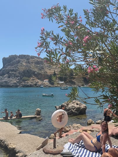 A wide shot captures a picturesque bay in Lindos, Greece, under a clear blue sky. In the foreground, a woman in a blue bikini sunbathes on a striped towel on a pebble beach. To her left, a white inflatable ring is held by a person partially visible, while another person in a black swimsuit stands further back. To the left, a wooden platform extends into the turquoise water, with a family of four, including two children, sitting on it. The water is calm, with a small white boat moored in the middle ground and other boats visible in the distance. A rocky, tree-covered hillside with a small white building and an orange roof dominates the background. Blooming oleander branches with pink flowers frame the scene from the right and upper part of the image.