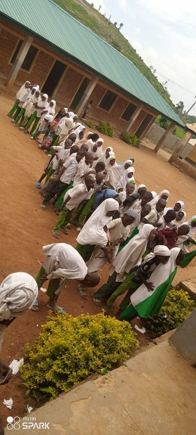 In Kagarko North, Nigeria, a group of schoolchildren, predominantly wearing white shirts and green pants, are lined up outside a brick building with a green corrugated metal roof. The building appears to be a school, with visible windows and pillars. The children, a mix of boys and girls, are mostly wearing hijabs or headscarves, with a few boys having short hair. Some children are looking at the camera, while others are engaged in conversation or looking down. A few children are holding bags. The ground is covered with reddish-brown dirt, and a yellow-green bush is visible in the foreground. The background shows a grassy hill with scattered trees, suggesting a rural or semi-rural setting. The sky is partly cloudy. The scene is captured from a slightly elevated angle.