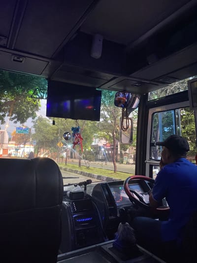 The image depicts the interior of a bus or large vehicle, viewed from a passenger's perspective looking towards the front windshield, at twilight. The scene is located on a road, identifiable through a visible blue directional sign as leading to "Cinunjul" and "Ciwidey" in West Java, Indonesia, consistent with the broader region of Parungserab.

In the foreground, the back of a black passenger seat is visible on the left. To the right, a male bus driver is seated, visible from the back, wearing a blue short-sleeved shirt and a dark baseball cap. His right hand is resting near his chin or jaw, and he is looking forward through the windshield. The driver's area includes a black steering wheel with a red trim, a dashboard with illuminated digital displays showing green and red indicators, and a gear stick with a fuzzy blue and white cover. Below the main dashboard, an audio system with blue illuminated controls is visible. A white tissue box or similar container rests on the dashboard.

Overhead, a flat-screen monitor is mounted to the ceiling, displaying dark colors with some faint reflections. A white air freshener dispenser is also mounted on the ceiling. Hanging from the central rearview mirror are a string of brown beads, a small disco ball ornament, and a red and blue character ornament. A smaller mirror is positioned below the main rearview mirror.

Through the windshield, an asphalt road with a curbed median strip is visible. Numerous green-leaved trees line both sides of the road and the median. In the background, low-rise buildings are visible, illuminated by various artificial lights, suggesting shops or residences. Streetlights along the road are also illuminated. A yellow diamond-shaped sign is partially visible on the left side of the road. The overall lighting indicates either early morning or late evening.