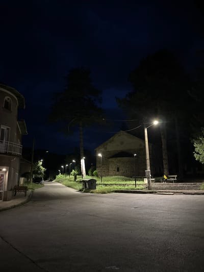 This is a nighttime, eye-level, medium shot of a street in Golyama Zhelyazna, Bulgaria. The sky is dark blue, indicating it is either twilight or a very dark night. On the left side of the frame, a building with a light-colored facade, arched windows, and a balcony is visible. A wooden bench is situated in front of this building. The street itself is paved and curves gently to the right, leading into the distance.
Along the right side of the street, a stone church with a light-colored, circular decorative element above its entrance is prominent. In front of the church, there are steps leading up to it. A wooden bench is placed on these steps, and a person wearing a bright yellow jacket is sitting on it. Several streetlights are illuminated along the street, casting pools of light on the pavement and surrounding greenery. Tall trees with dark foliage line the right side of the road, partially obscuring the view behind them. Two large, dark-colored trash bins are visible near the church. The overall atmosphere is quiet and serene, with the artificial lights creating a stark contrast against the dark night sky and surrounding shadows.