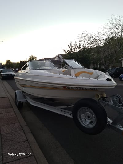 A white boat with orange and yellow accents is parked on a trailer on the side of a street in Woodburn, United States of America. The boat is a Bayliner, with the model number "5536-NU" visible on the side. The trailer has a spare tire mounted on it, and the text "EZ LOADER" is visible on the frame of the trailer. The boat is facing towards the right side of the image. In the background, there are houses and trees. A black car is parked on the left side of the street, and another car is visible in the distance. The sky is clear and the sun is setting. The image was taken at dusk.