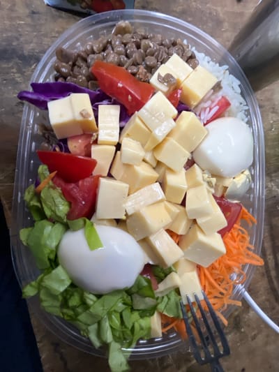 A close-up, elevated shot shows a colorful salad packed in a clear plastic, oval-shaped container, resting on a textured wooden surface. The salad is composed of various ingredients neatly arranged in separate sections.
Dominating the center are numerous light-yellow cheese cubes. To the left, a large portion of brown lentils is visible, alongside some white rice mixed with small red tomato pieces and purple cabbage. On the right side, a generous amount of vibrant orange shredded carrots is present, next to a bed of chopped green lettuce. Two whole, peeled hard-boiled eggs are positioned in the container; one is prominent on the left, and another is partially visible on the right, nestled amongst the lettuce and carrots. A black plastic fork is partly submerged in the cheese and carrots on the top right, suggesting the meal is being eaten or is ready to be consumed. Another white plastic fork is discernible in the upper background.
The lighting appears bright, highlighting the fresh appearance of the ingredients. There is no visible text within the image. This meal is likely a pre-packaged or take-away option, consistent with a quick and convenient meal. The setting appears to be an indoor environment, possibly a dining table. The location context provided is Campana, Argentina.