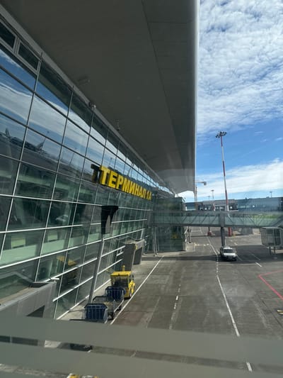 This image displays the exterior of Terminal 1A at Kazan International Airport (KZN) in Stolbishche, Russia. The modern building features a multi-story glass facade that reflects a partially cloudy blue sky and distorted outlines of an aircraft. Large yellow Cyrillic lettering, "ТЕРМИНАЛ 1А" (Terminal 1A), is affixed to the glass. In the foreground on the asphalt tarmac, a yellow ground service vehicle, possibly a baggage tug, is visible, connected to a train of three blue-framed baggage carts. Further along the tarmac, a white utility vehicle with blue markings is parked. A skybridge, featuring a glass-enclosed walkway, traverses the tarmac area, connecting different sections of the airport infrastructure. A tall, slender light pole, striped red and white, with a cluster of floodlights at its apex, is positioned on the right side of the frame. The tarmac is marked with white solid and dashed lines, and red lines. The sky is partly cloudy. No individuals are visible.