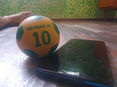 A slightly out-of-focus, eye-level medium shot shows a yellow toy soccer ball with green accents and the text "NEYMAR JR 10" in green lettering, resting on a wooden floor. To the right of the ball, a black smartphone lies face up. In the background, a portion of a person's bare foot is visible on the left, and a wall with a green, textured pattern is on the right. The scene suggests an indoor domestic setting. The image was taken in Vahdat, Tajikistan.