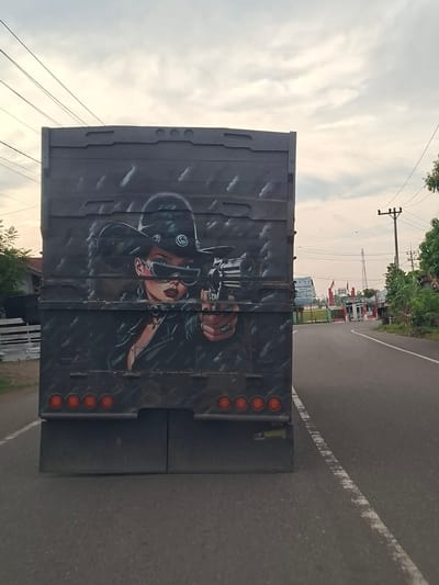 The image shows the back of a flatbed truck driving on a paved road, likely within Mee Pangwa, Indonesia, based on the surrounding environment and context. The truck bed is painted with an image of a woman holding a revolver. Traffic lights are visible at the back of the truck. Overhead power lines run across the scene, and in the background, there are buildings and trees. The road surface is grey asphalt, with a visible white line marking the lane.